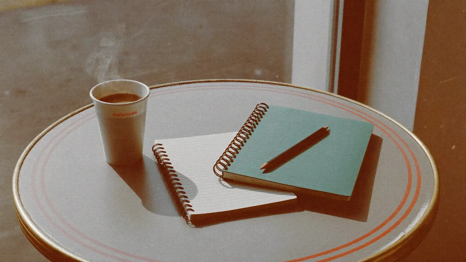 A coffee cup, a spiral notebook and a pencil on a café table by a sunny window