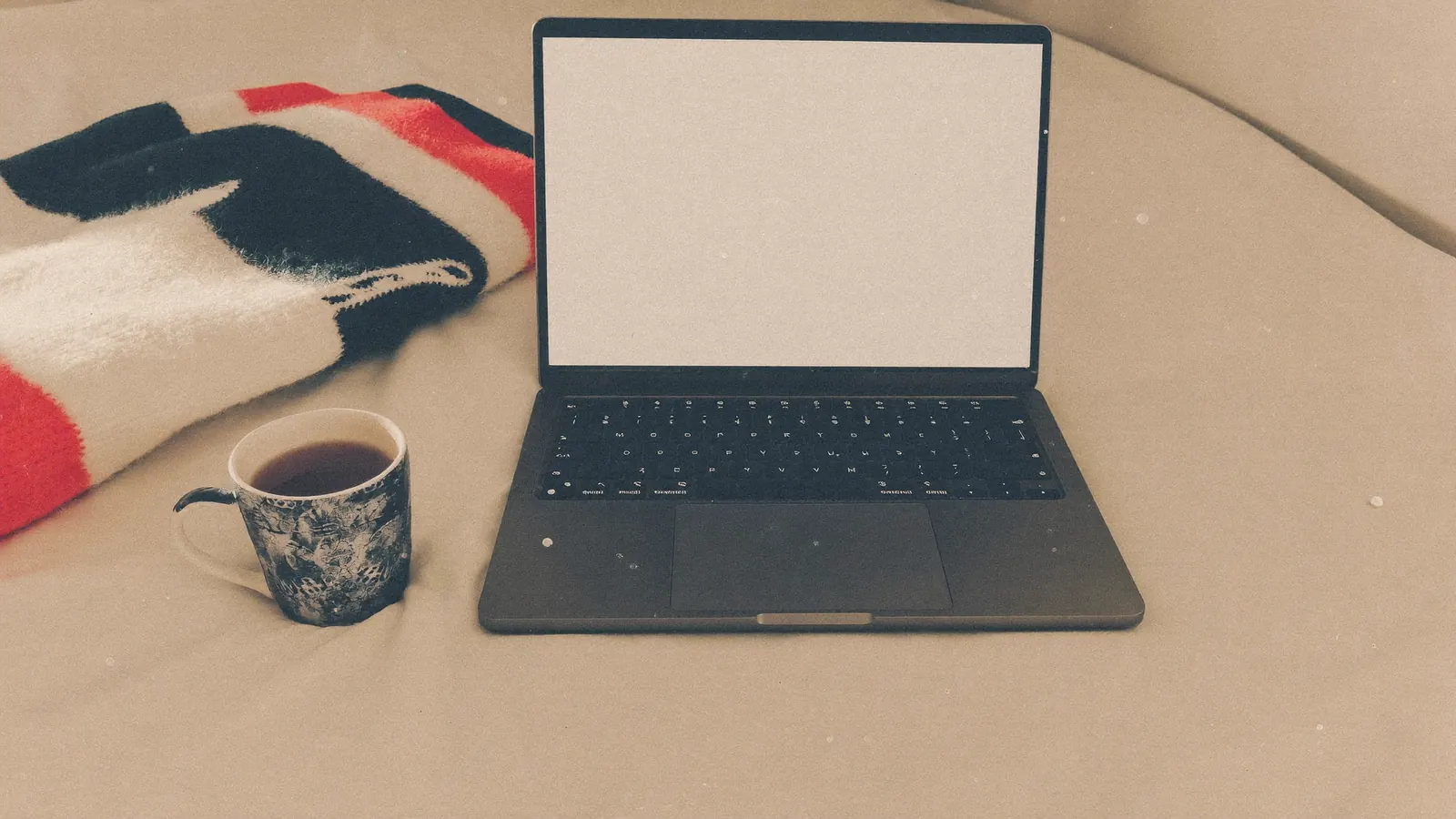 A laptop on an unmade bed with a mug of tea and a folded cardigan, morning light