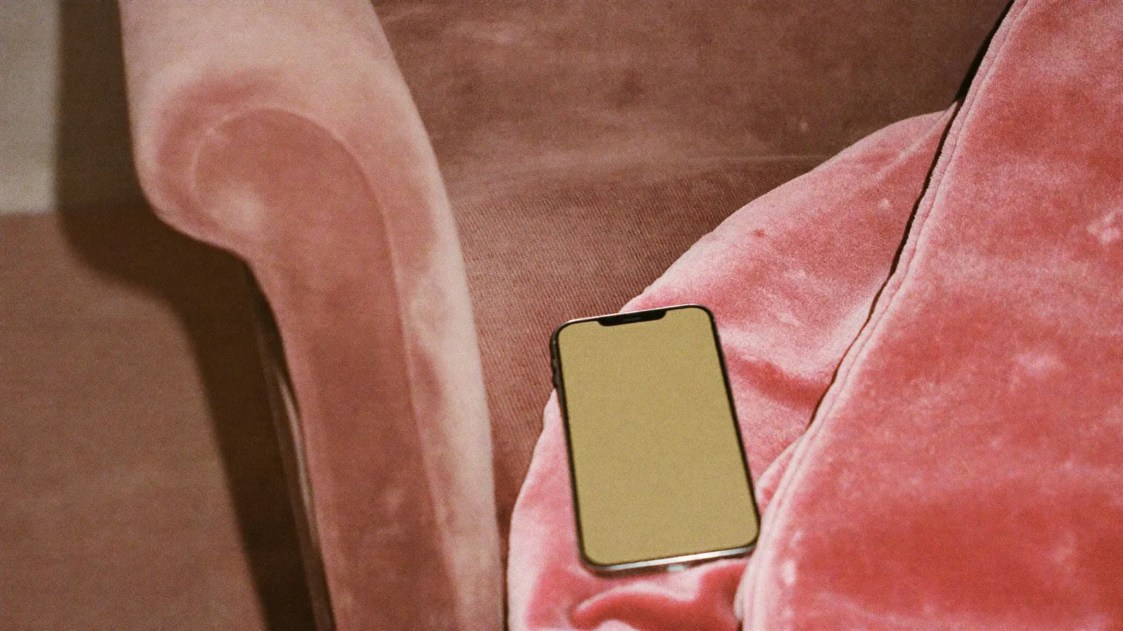 A smartphone face up on a velvet armchair with a soft pink throw