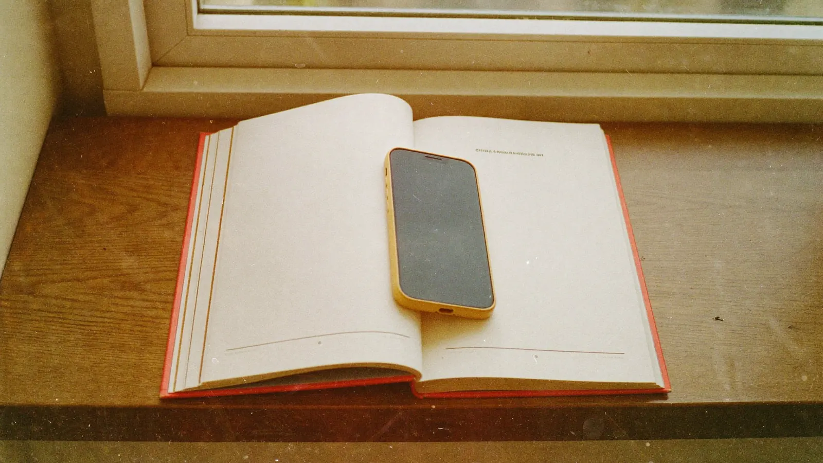 A smartphone resting on an open paperback book on a wooden windowsill in golden light