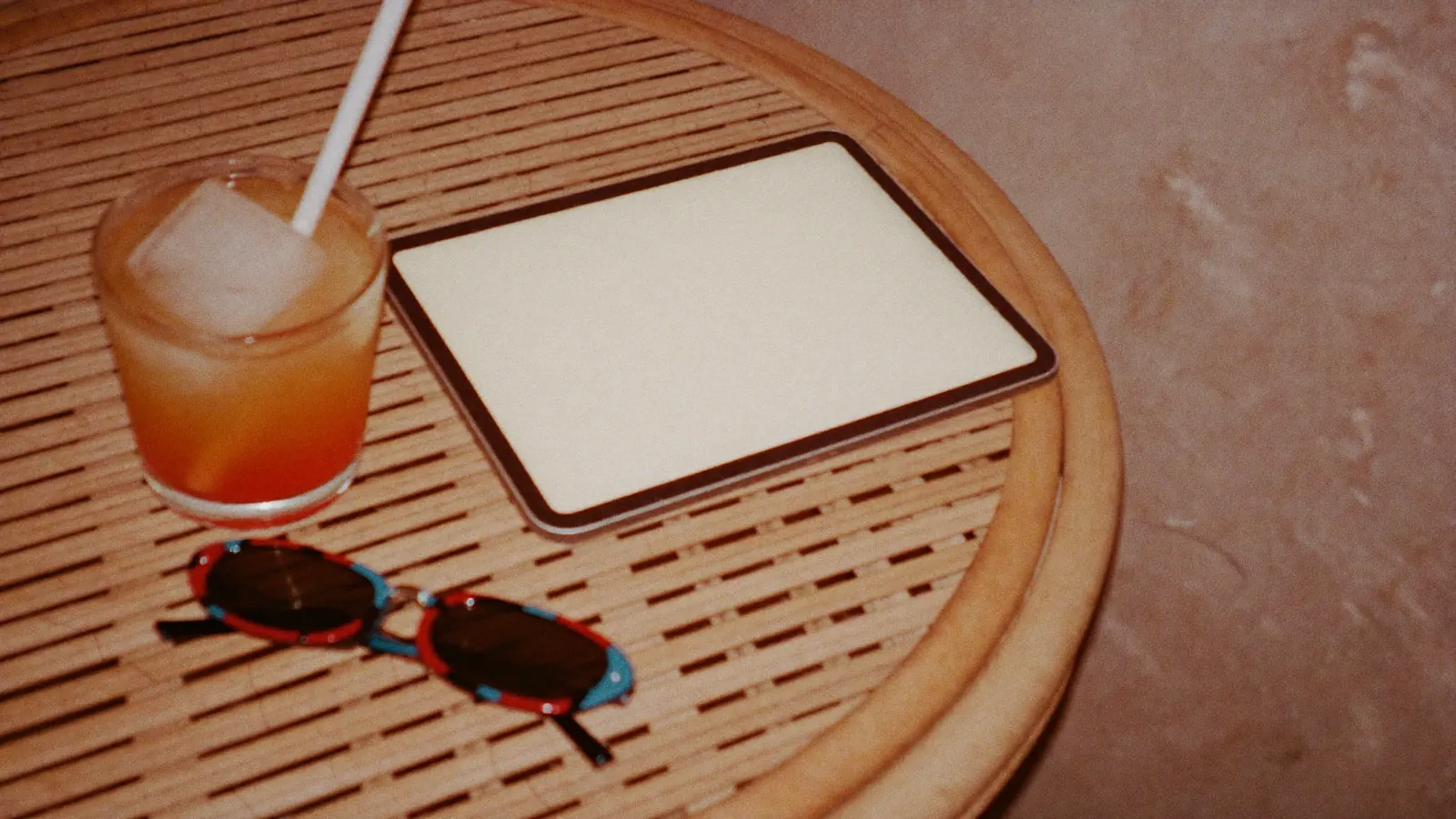 A tablet on a rattan side table beside a glass of iced tea and sunglasses