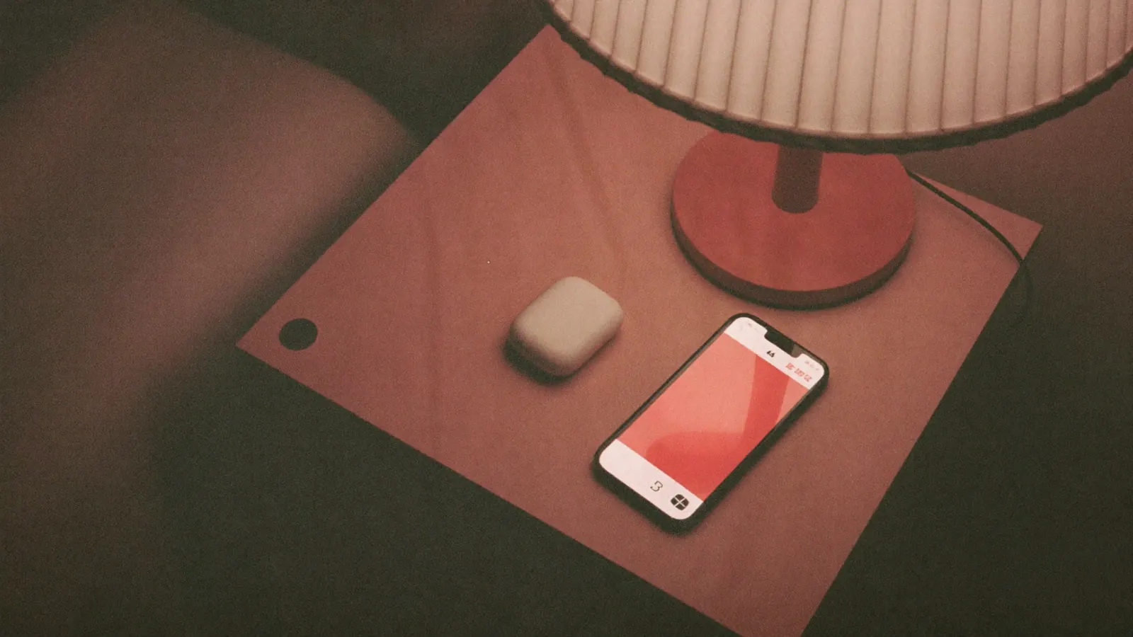 Wireless earbuds case and a phone on a bedside table next to a dim lamp